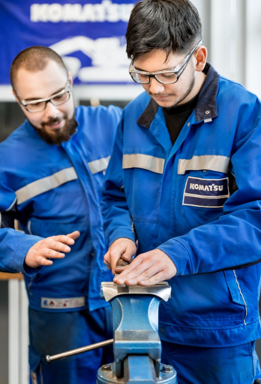 Zwei Männer in Komatsu-Arbeitskleidung bei der Metallbearbeitung an einem Schraubstock