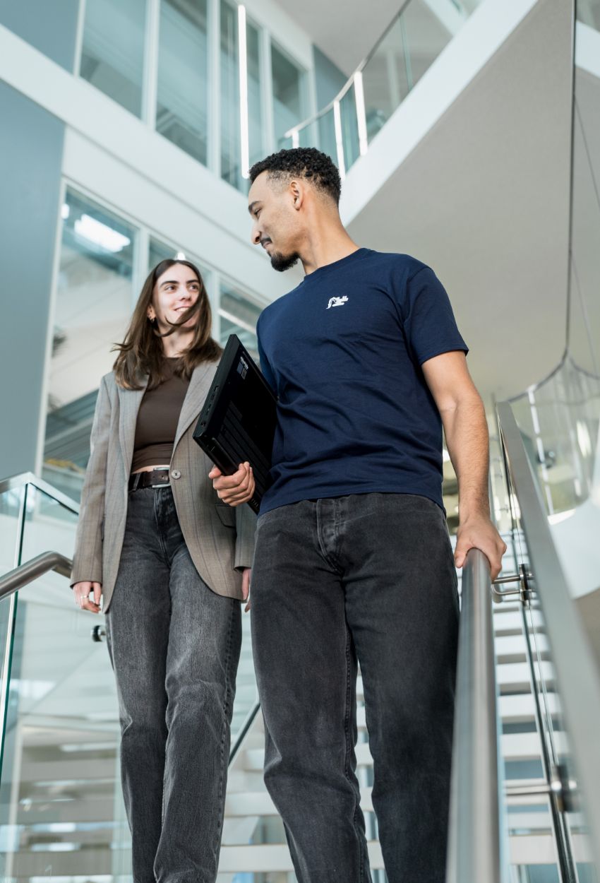 Zwei junge Erwachsene gehen eine Treppe in einem modernen Bürogebäude hinunter, der Mann trägt ein dunkelblaues T-Shirt und hält einen Laptop, die Frau trägt ein graues Sakko und dunkle Jeans